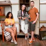 Three people relax on a porch beside a camper, enjoying a casual moment together in a serene outdoor setting